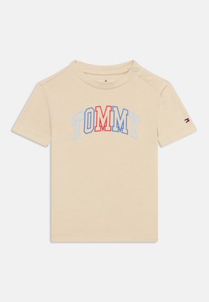 T-shirt beige à manches courtes en coton, avec l'inscription "TOMMY" en lettres arquées rouges et bleues sur le devant. Détail à boutons-pression sur l'épaule.
