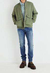 Olivgrön vadderad jacka med rund krage, kombinerad med en randig skjorta, blå jeans och bruna snörkängor. Har framfickor.