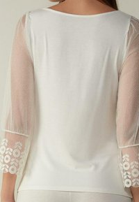 Top bianco con maniche in tulle trasparente a tre quarti con texture, decorate con ricami floreali ai polsi. Tessuto morbido e vestibilità comoda.