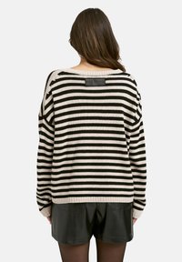 Eine Frau trägt einen schwarz-beigen, horizontal gestreiften Pullover und schwarze Shorts, von hinten betrachtet vor einem einfarbigen Hintergrund.