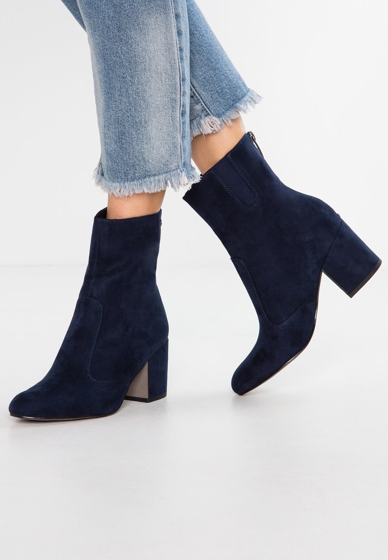 Femme portant des bottines en daim bleu foncé à talons épais et des jeans à ourlets effilochés et délavés, debout sur un sol blanc avec un fond blanc.