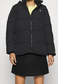 Czarna kurtka puchowa z kapturem, z zamkiem z przodu, pikowanym wzorem i bocznymi kieszeniami, noszona na czarną spódnicę.