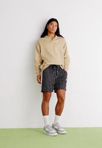 Beżowa koszulka polo z kołnierzykiem, czarno-białe spodenki w paski, białe skarpetki i szare sneakersy. Model stoi na tle gładkiej ściany.