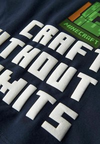 Jūras zils T-krekls ar teksturētām baltām burtiem, kas rakstīts "RAŽO BEZ TRIECIENIEM" un Minecraft logo zaļā un oranžā krāsā.