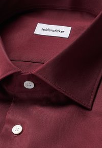 Camicia borgogna con una texture liscia e satinata, dotata di un colletto classico e due bottoni bianchi vicino al colletto. L'etichetta reca la scritta "seidensticker."