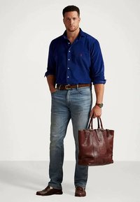 Mężczyzna w niebieskiej koszuli z guzikami i dżinsach trzyma brązową skórzaną torbę, stojąc na jednolitym tle.