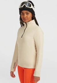 Beige fleece-pusero, jossa on puolivetoketju kauluksessa ja Polartec-brändäys vasemmassa hihassa. Yhdistetty kirkkaan oransseihin alaosiin.