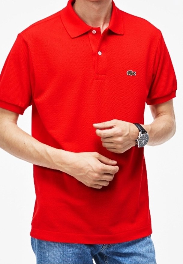 Homme portant un polo rouge vif à manches courtes avec un petit logo crocodile sur la poitrine et une montre noire au poignet gauche.