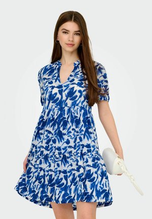 Blomstret blå kjole med korte pufærmer, nøglehulskræft, lagdelt nederdel og let tekstur; parret med en hvid fletpet. 