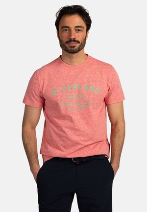 Man met kort donker haar en baard, die een rood T-shirt draagt met de tekst "N-ZEALAND" en coördinaten, met handen in de zakken, tegen een effen achtergrond.