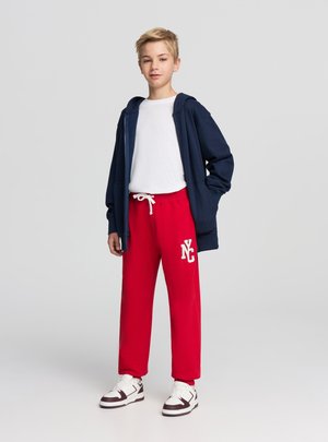 Felpa con cappuccio blu navy con zip sopra una t-shirt bianca, abbinata a pantaloni della tuta rossi con un logo bianco, completata da sneakers bianche.