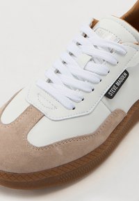 Sneaker in pelle bianca e suede beige con lacci bianchi, suola in gomma marrone e un'etichetta nera con la scritta "STEVE MADDEN" di lato.
