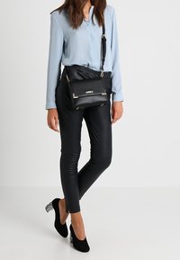 Černá crossbody taška se zlatými detaily, obdélníkový tvar, hladká kožená textura. Kombinováno s upnutými černými kalhotami a světlou modrou blůzou.