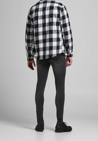 Hombre vistiendo una camisa a cuadros blanco y negro, jeans ajustados gris oscuro y zapatos de cuero negros, de pie contra un fondo gris liso.