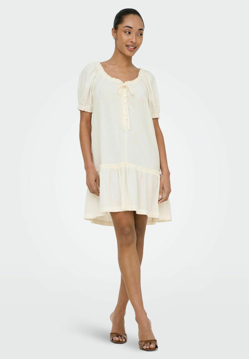 Femme debout, portant une robe courte et ample crème avec manches bouffantes et boutons à l'avant, associée à des talons ouverts.
