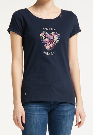 Granatowa koszulka z krótkimi, podwiniętymi rękawami, z graficznym sercem w kwiaty, tekst "Słodkie Serce", noszona z niebieskimi dżinsami przez stojącą osobę.