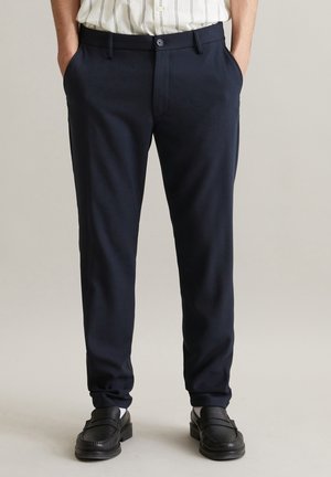 Man draagt marineblauwe, op maat gemaakte broek met zijzakken, zwarte loafers en een wit overhemd met dunne verticale strepen, ingestopt.