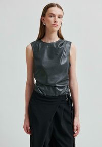 Žena s dlouhými hnědými vlasy, oblečen�á v bezrukávovém černém koženém topu a v černé sukně s vysokým pasem a bočním přezkou na jednofonním pozadí.