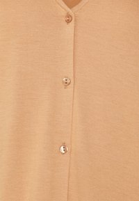 Beige neuletakki, jossa on V-pääntie, viisi nappia edessä ja sileä, tekstuuripintainen kangas. Yksinkertainen muotoilu ilman kuvioita tai yksityiskohtia.