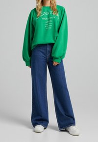 Zelená oversized mikina s bílým potiskem, zkombinovaná s vysokými modrými volnými džíny a bílými teniskami. Hladký materiál a pohodlný střih.