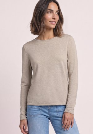 Femme aux cheveux bruns mi-longs, portant un haut beige à manches longues et un jean bleu clair, regardant légèrement sur le côté avec un sourire.