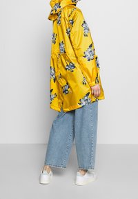 Chaqueta impermeable amarilla con estampado floral en azul y blanco, que presenta una capucha, puños elásticos y un corte relajado. Lleva puesta sobre unos jeans sueltos azules.