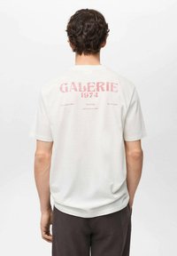Bílé bavlněné tričko s potiskem "GALERIE 1974" v bledě růžové barvě na zadní straně. Krátké rukávy, kulatý výstřih a volný střih.