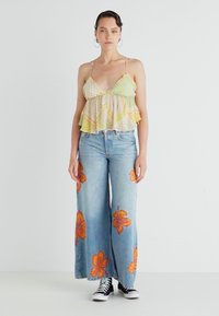 Lys grønn og gul mønstret rufsete topp med spaghettistropper, brukt med vide falmede blå jeans med oransje hibiskusblomstertrykk.