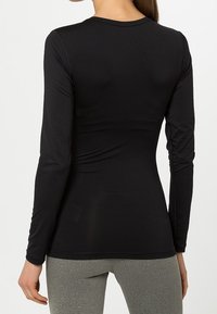 Žena oblečená v černém přiléhavém tričku s dlouhým rukávem a šedých legínách, zobrazená zezadu na jednobarevném pozadí.