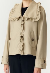 Veste beige avec bordure froncée sur le col et le devant, poignets boutonnés et boutons marron sur les poches, portée sur un haut noir.