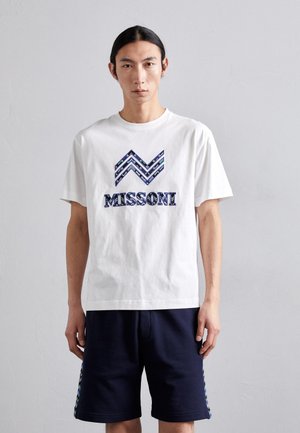 T-shirt en coton blanc présentant un motif en zigzag bleu et "MISSONI" en lettres majuscules, associé à un short bleu marine et des accents texturés.