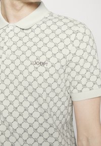 Polo beige avec un subtil motif floral géométrique et le logo "JOOP!" brodé sur la poitrine, porté par une personne.
