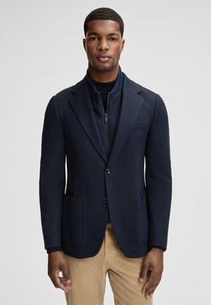 Marineblazer met twee opgestikte zakken, sluiting met één knop en een zachte textuur. Gelaagd over een donkerder coltrui.