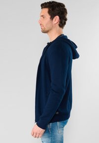 Marineblauwe hoodie met ritssluiting en een capuchon, gemaakt van zachte stof. Heeft een gespleten kangoeroezak en ribgebreide boorden. Draagt over lichtblauwe jeans.
