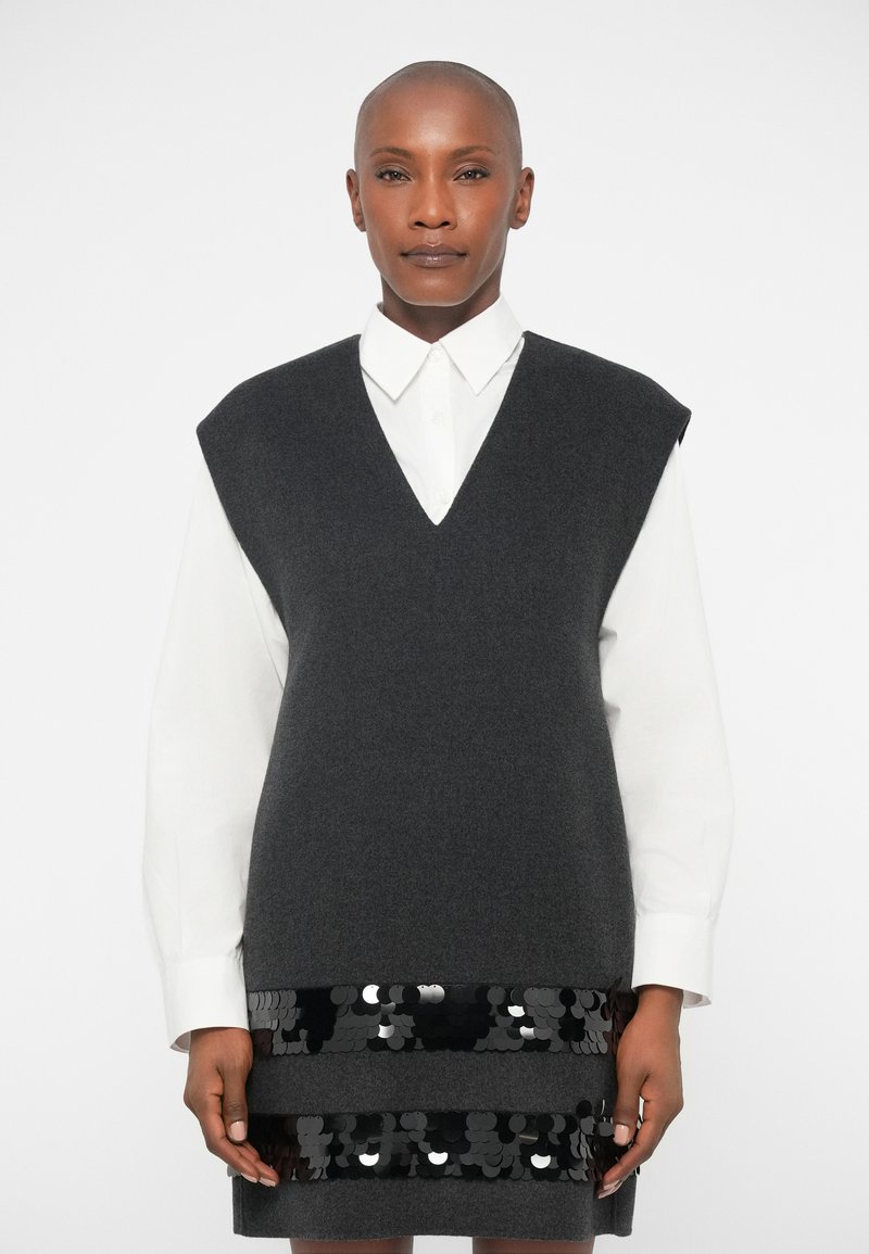 Um colete de malha cinza sem mangas em tecido suave, sobre uma camisa branca com colarinho. Apresenta um decote em V e um detalhe texturizado de lantejoulas pretas na parte inferior.