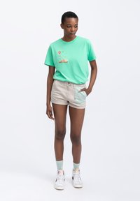 T-shirt in cotone verde menta con design stampato, pantaloni corti beige con tasca azzurro chiaro, sneakers bianche e calzini verde menta. Outfit estivo informale.