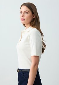Witte polo met korte mouwen en een gestructureerd breipatroon, drie-knoopsluiting en een zachte kraag. Draagt met een donkerblauwe broek en een riem.