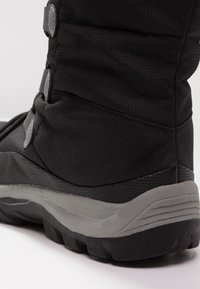 Botte d'hiver noire avec tige en textile matelassé, semelle en caoutchouc grise et accents de boutons. Présente un design texturé et un matériau durable.