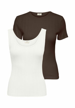 Jeune femme souriante, debout les mains dans le dos, portant un haut marron à manches courtes, un pantalon noir à jambes larges et des chaussures blanches pointues.