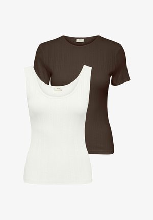 Jeune femme souriante, debout les mains dans le dos, portant un haut marron à manches courtes, un pantalon noir à jambes larges et des chaussures blanches pointues.
