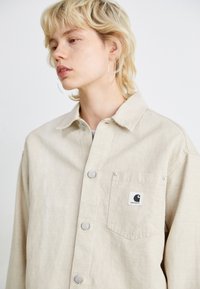 Beige button-up jacket made of cotton, featuring a chest pocket with a logo, silver buttons, and a relaxed fit design.