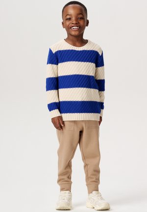 Blau- und cremefarbener gestreifter Strickpullover mit strukturierter Oberfläche, kombiniert mit beigefarbenen Jogginghosen und weißen Sneakers.