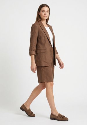 Vrouw lopend in een bruine blazer en bijpassende knielange korte broek met een wit topje en bruine loafers met kwastjes op een witte achtergrond.