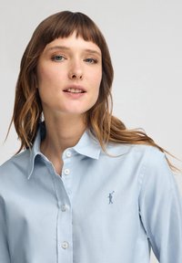 Ljusblå knappskjorta i bomull med enfärgad textur. Har en liten broderad logotyp på vänster bröst och en klassisk krage.