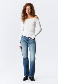 Kvinne med krøllete hår iført en hvit off-shoulder langermet topp, blå jeans og svarte sko, står med den ene hånden i lommen.