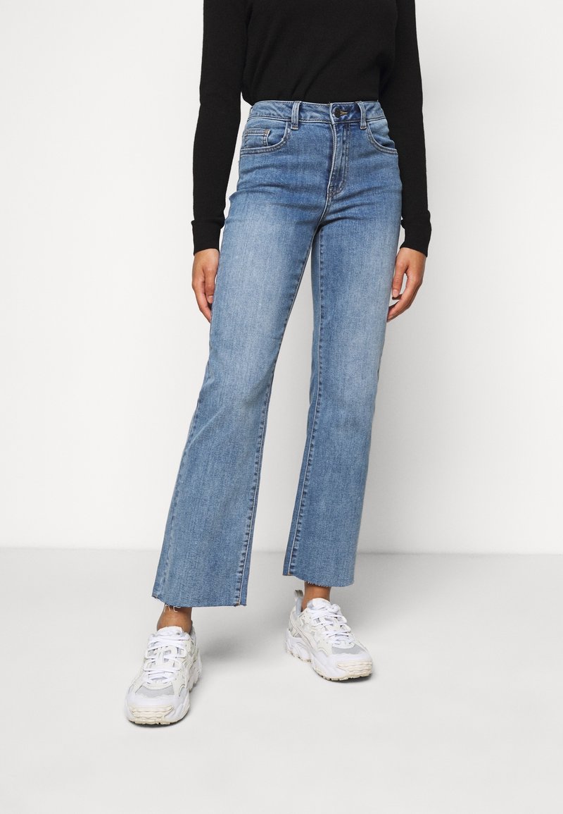 Jean large bleu clair à taille haute, avec un design traditionnel à cinq poches et des ourlets effilochés, associés à des baskets blanches.