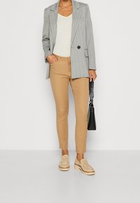 Blazer gris à rayures, porté sur un haut crème, associé à un pantalon slim beige et des mocassins beiges. Un sac bandoulière noir ajoute du contraste.