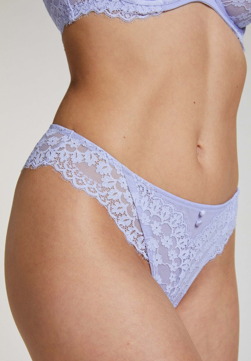 Lila kanten strings met scalloped randen en een contrasterende gladde tailleband. Voorzien van decoratieve knopen op de achterkant voor extra detail.