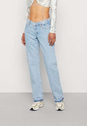 Person iført lyseblå højtaljede jeans med lige ben, en hvid cropped top med slangeskindsprint og hvide sneakers, stående mod en ensfarvet baggrund.