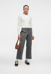 Žena stojí a na sebe má biely pletený kardigán, šedé široké nohavice cargo, čierne tenisky a svetlé ponožky. Drží malú hnedú kabelku.